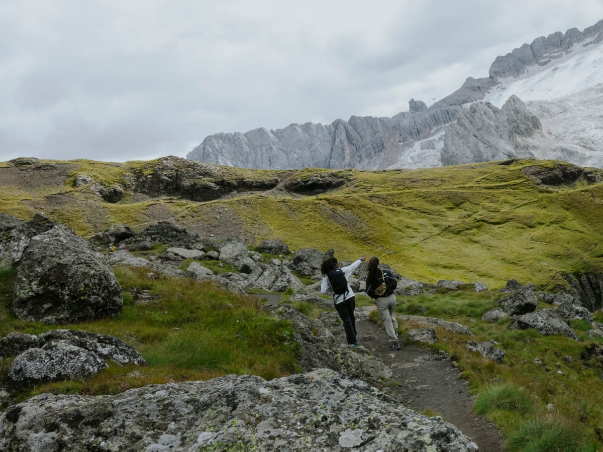 Two hikers moving along a rocky mountain path surrounded by green alpine landscape and rugged peaks.